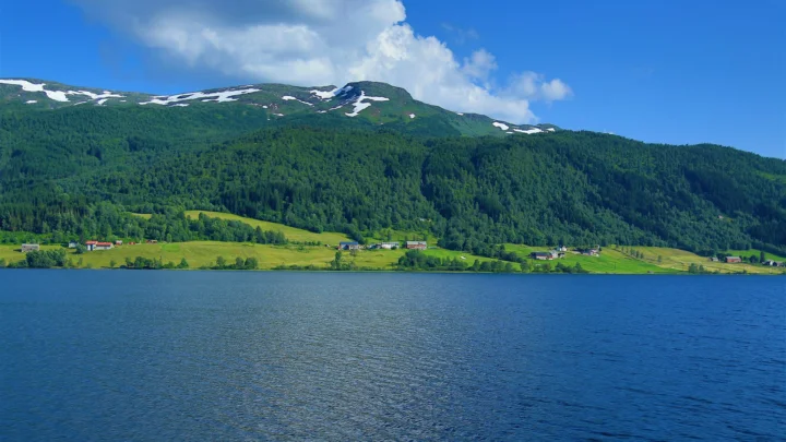 蓝色湖泊前坐落着森林覆盖的山脉，山顶残留着白雪，天空中飘着云朵。