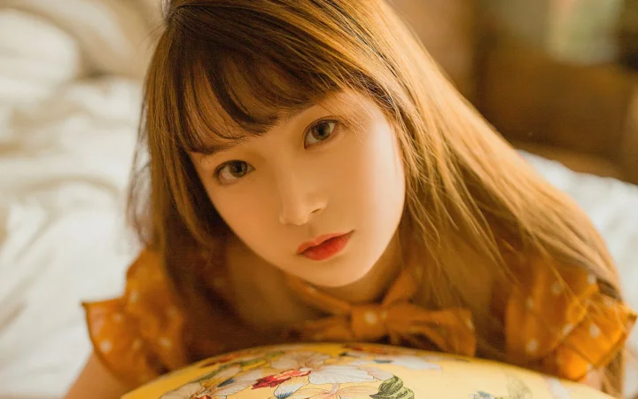 一名棕色长发女性在暖光下直视镜头，面前可见带图案的靠垫。