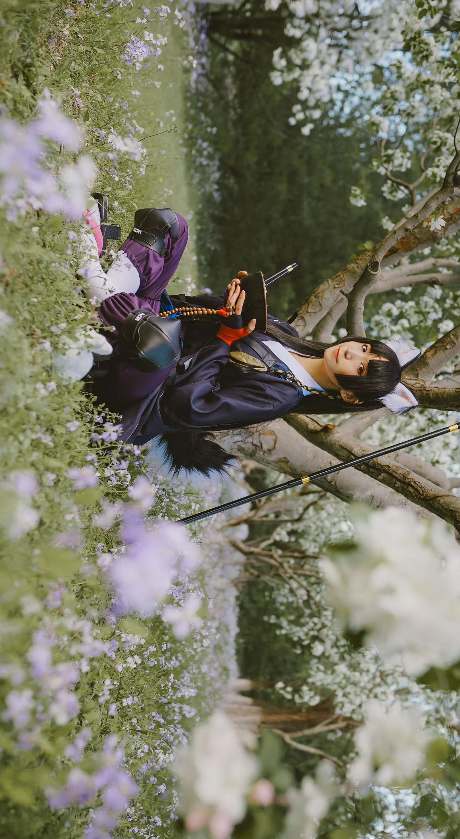暗い衣装のコスプレイヤーが、紫色の花が咲く木々の間に横たわっています。