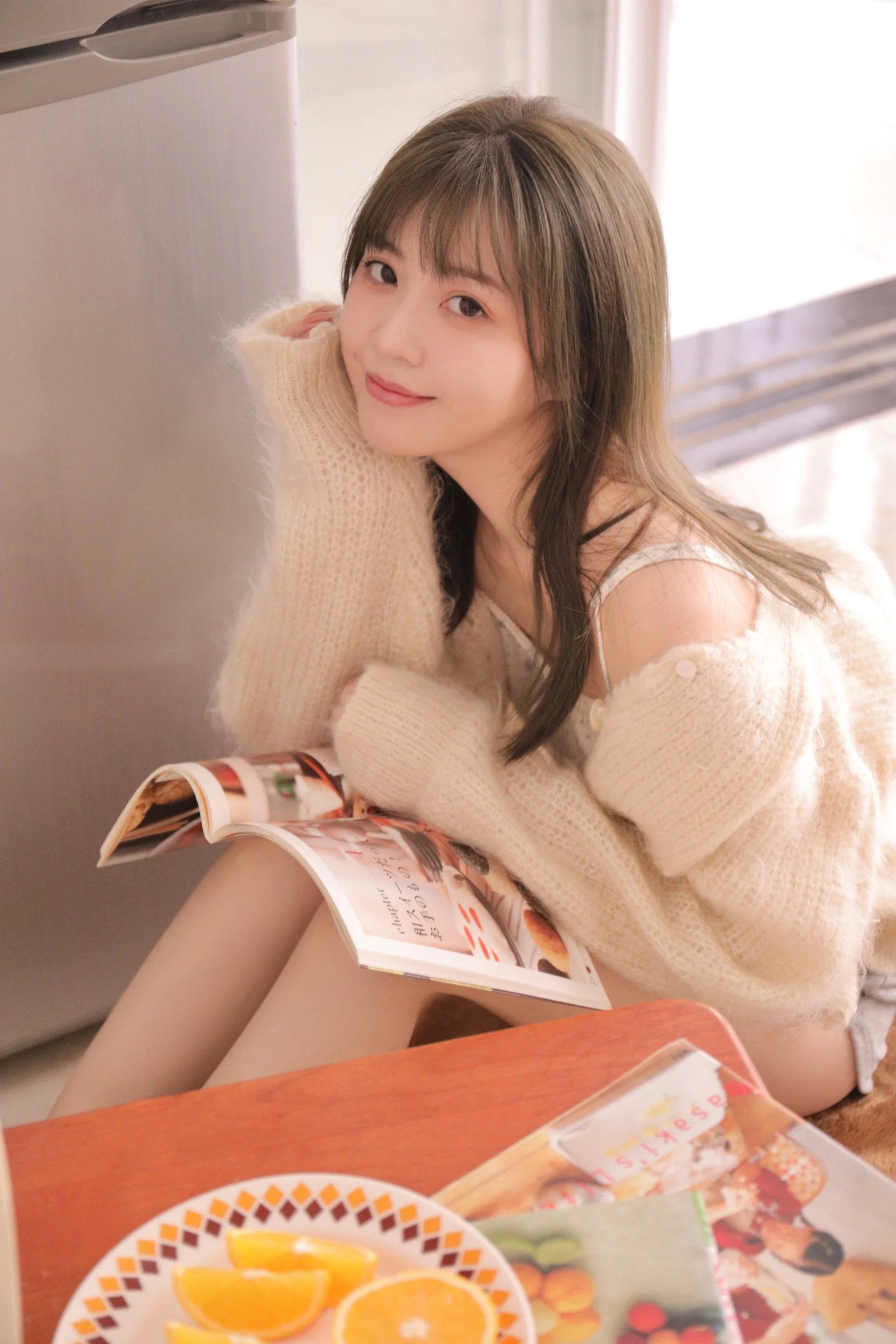 一名長髮女子穿著米色針織衫坐在室內，身旁放著雜誌和橙子。