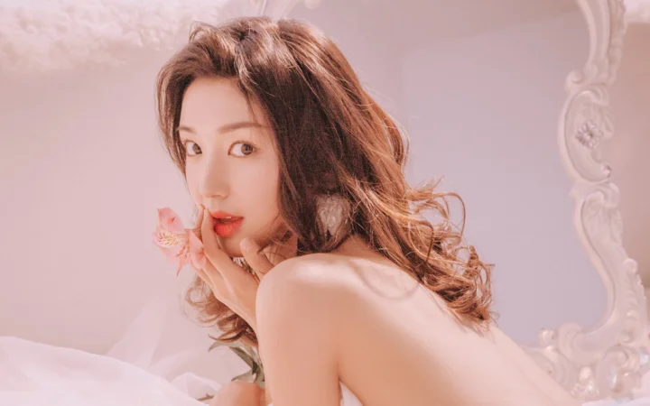 一位长卷发女性在柔和的粉色背景前回眸凝视镜头。