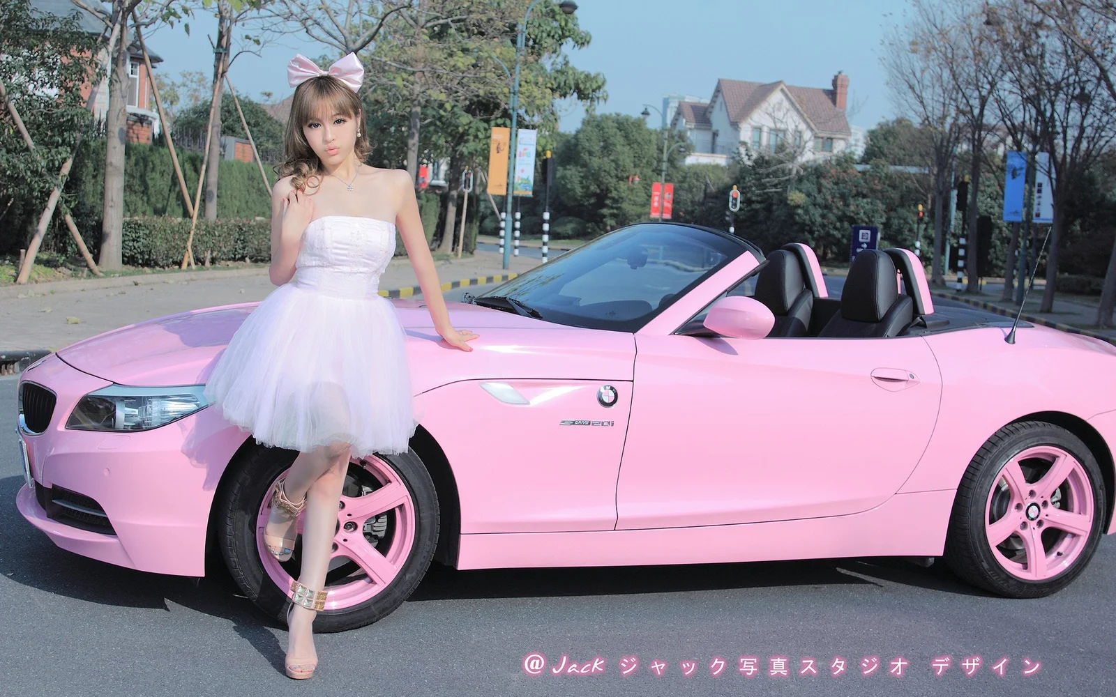 ピンクのリボンを着けた女性が、屋外の道路脇でピンクのオープンカーの前に立っています。
