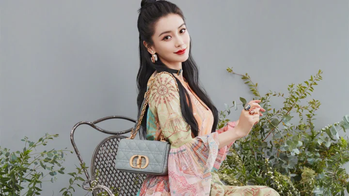 一位成年女性坐在自行车旁，手提灰色手袋，身后是灰色墙面与绿植。