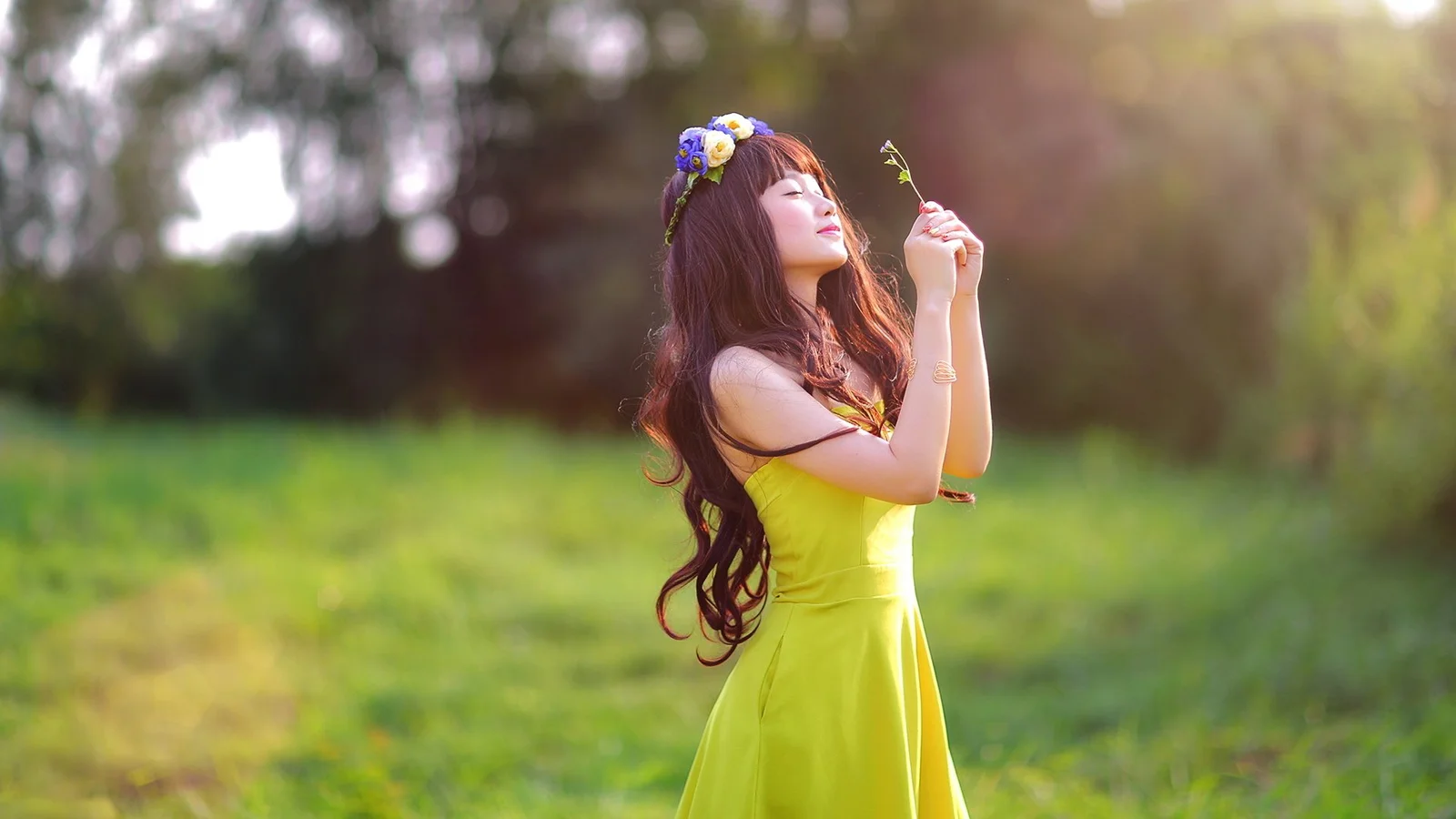 一名身穿黄色连衣裙的女子站在阳光洒落的草地上，手中拿着一朵小花。