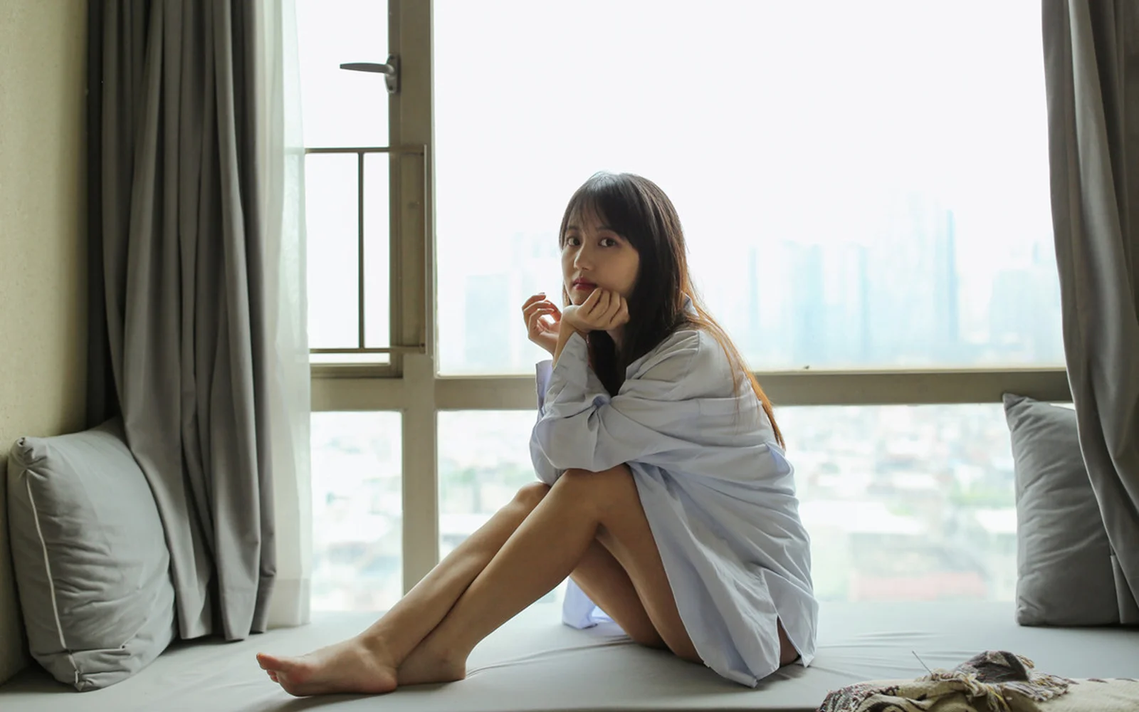 부드러운 실내 조명 속에서 성인 여성이 창가의 침대에 앉아 있고 창밖으로 도시 풍경이 보입니다.