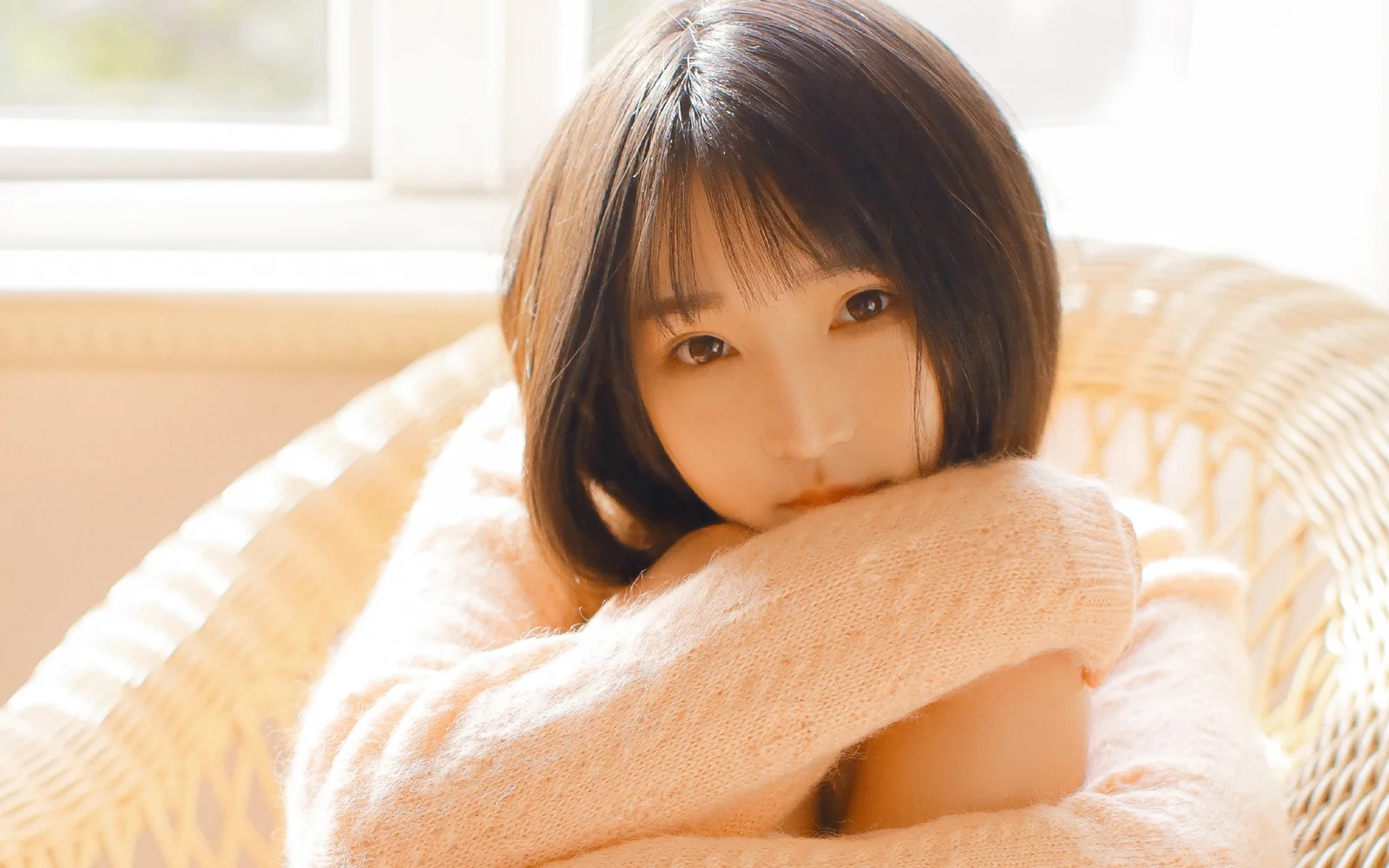 一名成年女性穿着浅色毛衣坐在藤椅上，在明亮的室内光线下望向镜头。