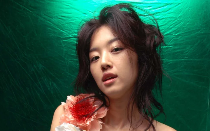 초록색 배경 앞에서 어깨 곁에 붉은색과 분홍색 꽃을 장식한 성인 여성이 카메라를 바라보고 있다.