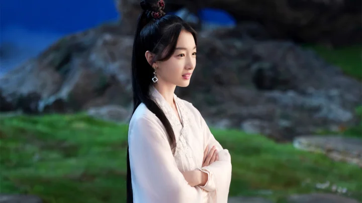 밝은 전통 의상을 입은 여성이 푸른 잔디와 바위를 배경으로 야외에 서 있다.