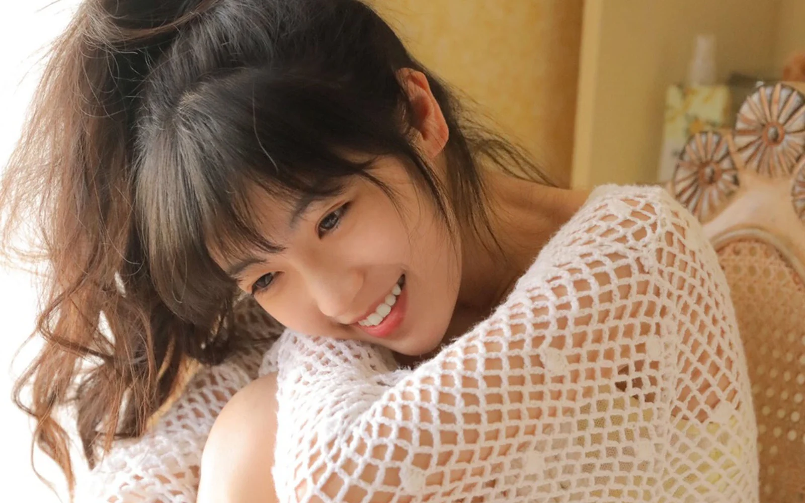 흰색 니트 상의를 입은 미소 짓는 여성이 실내에서 의자 등받이에 기대고 있습니다.