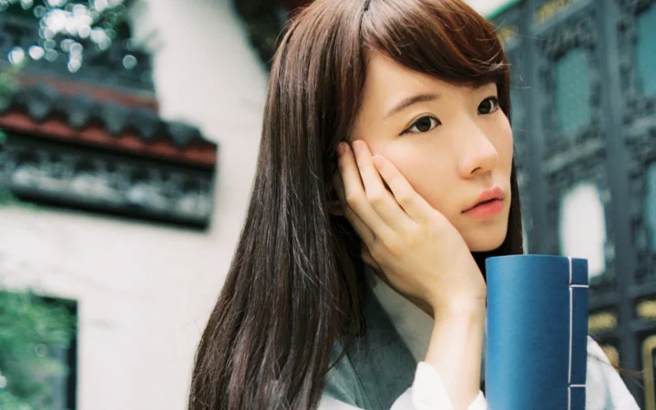 一名长发女子用手托着脸，身旁放着一本蓝色书籍，背景是虚化的建筑。
