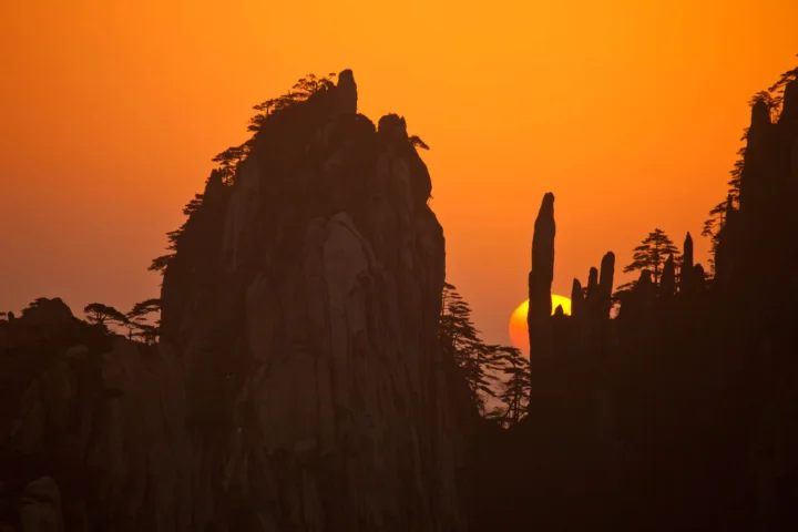 橙色夕阳映照着层叠的奇峰与松树剪影。