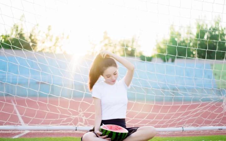 一名年轻女子坐在运动场的球门网前，手持切开的西瓜。