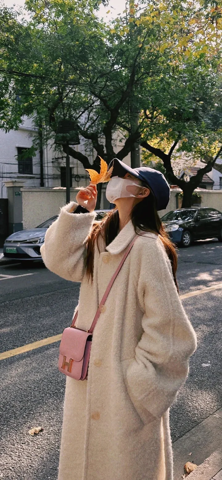 一名戴帽子的女子穿着米色长外套站在树荫下的街道旁。