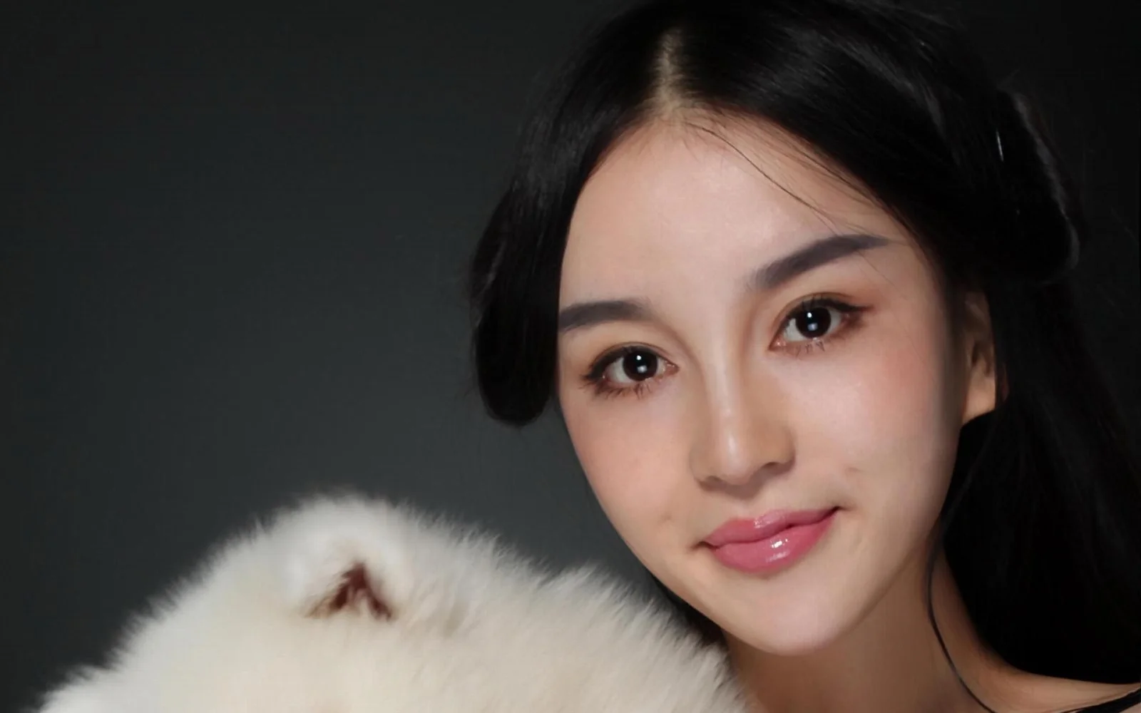 어두운 배경 앞에서 성인 여성이 하얗고 복슬복슬한 강아지와 함께 미소 짓고 있다.