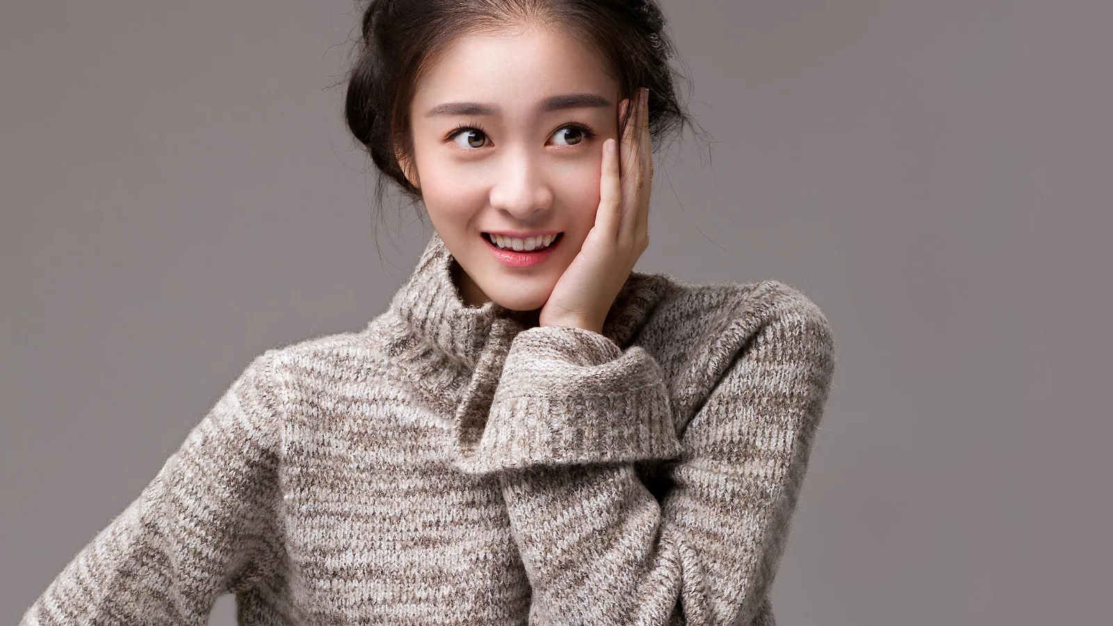 一名成年女子身穿灰色针织衫，在灰色背景前微笑并用手托着脸颊。