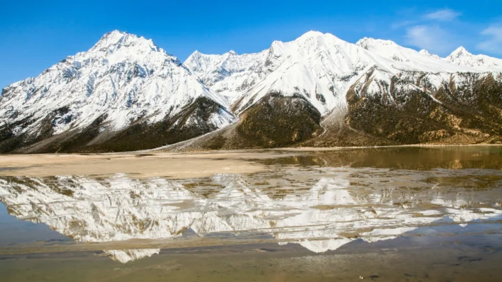 雪覆盖的群山映照在平静的湖面上，远处是晴朗的蓝天。