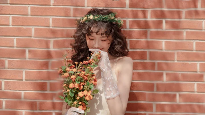 一名戴着花环、手捧花束的卷发女子站在红砖墙前。