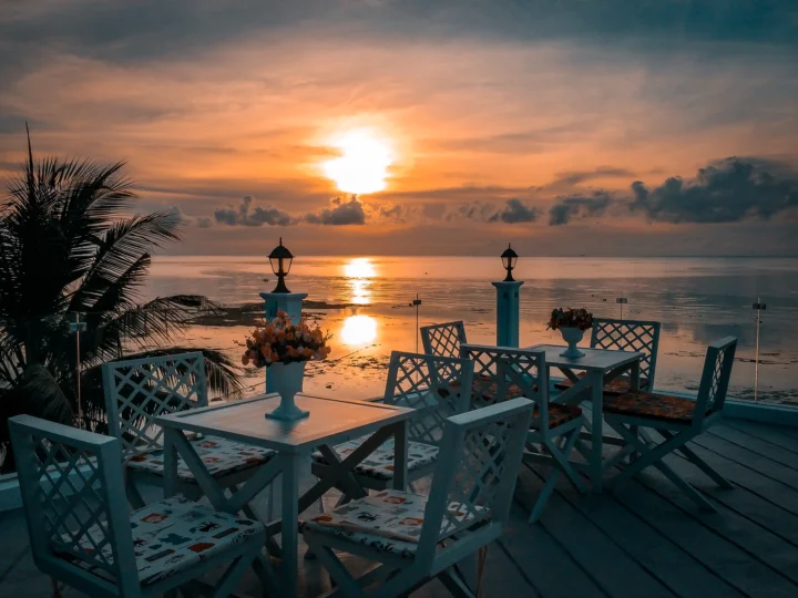 夕阳映照着平静的海面，前景是摆放在木质露台上的白色餐桌椅和棕榈树。