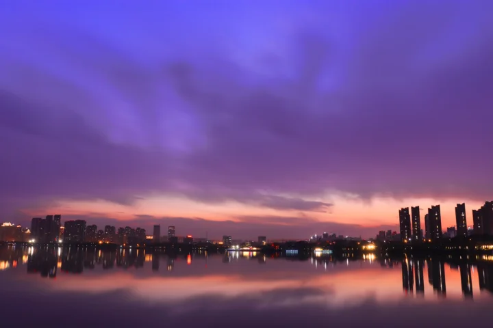 暮色天空下的城市天际线倒映在平静湖面上，湖岸灯光清晰可见。