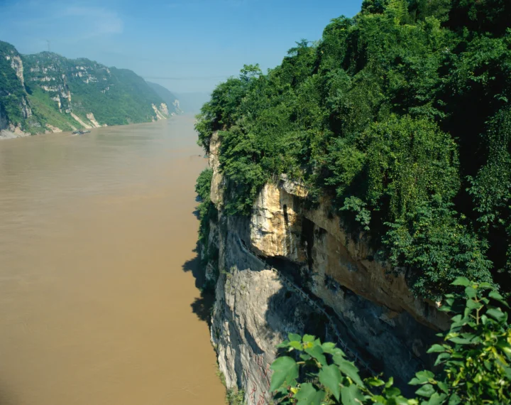 宽阔的浑浊河流穿过群山峡谷，近处是覆盖植被的陡峭岩壁。