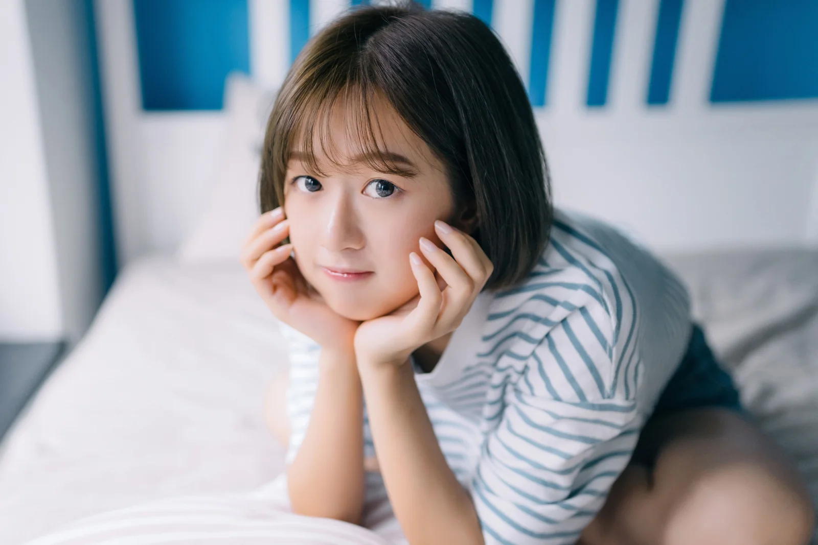 단발머리 여성이 파란색과 흰색 줄무늬 셔츠를 입고 실내 침대에서 두 손으로 턱을 괴고 있습니다.