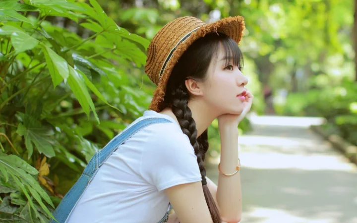 一名戴草帽并编着长辫子的女子坐在绿意盎然的林间小路旁。