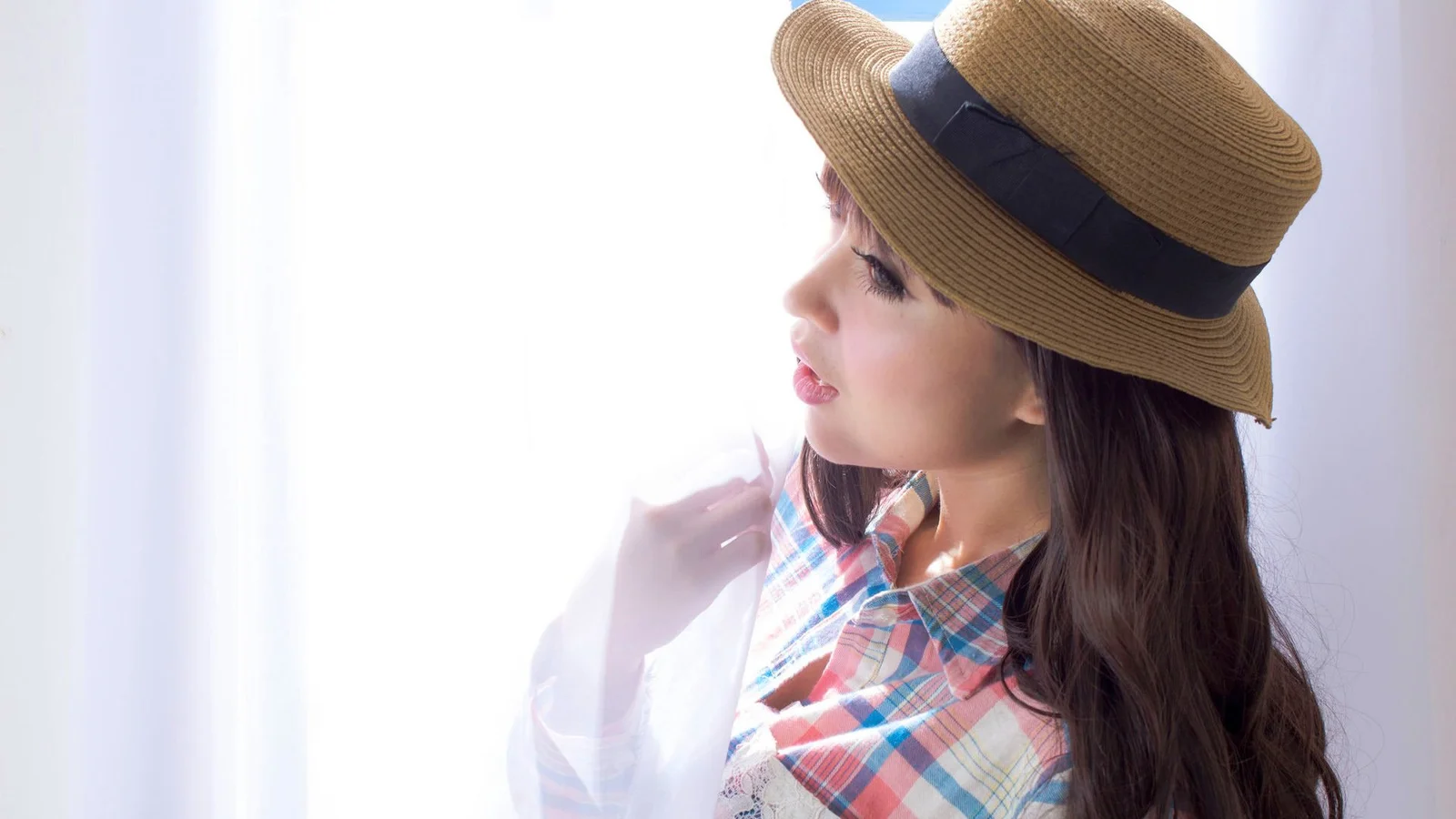 一名戴草帽、穿格纹上衣的女性靠近明亮窗边侧身望向一侧。