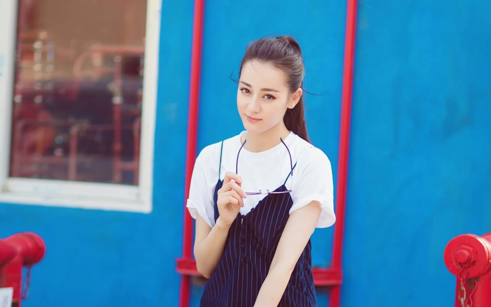 ポニーテールの女性が青い壁の前に立ち、眼鏡を手に持ち、白いトップスと濃色のストライプ柄ジャンパースカートを着ています。