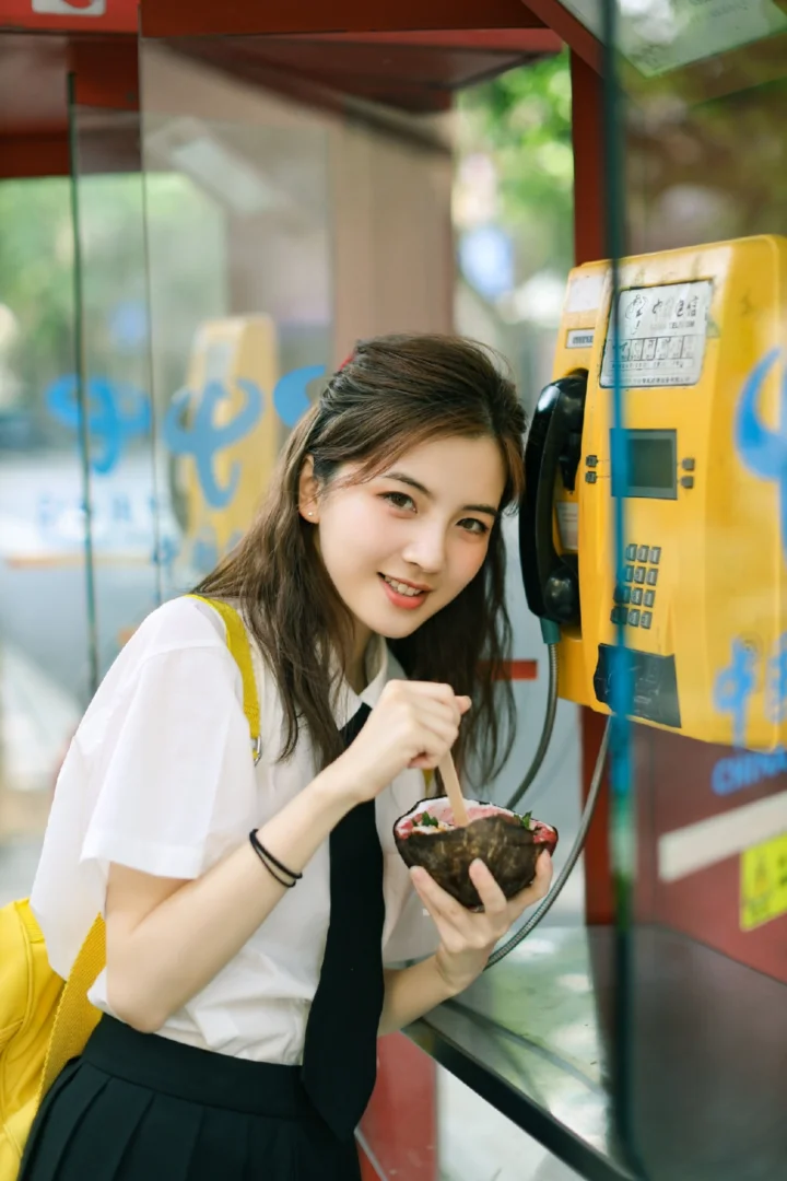 一名女子站在黄色公用电话旁，手里拿着碗和勺子。