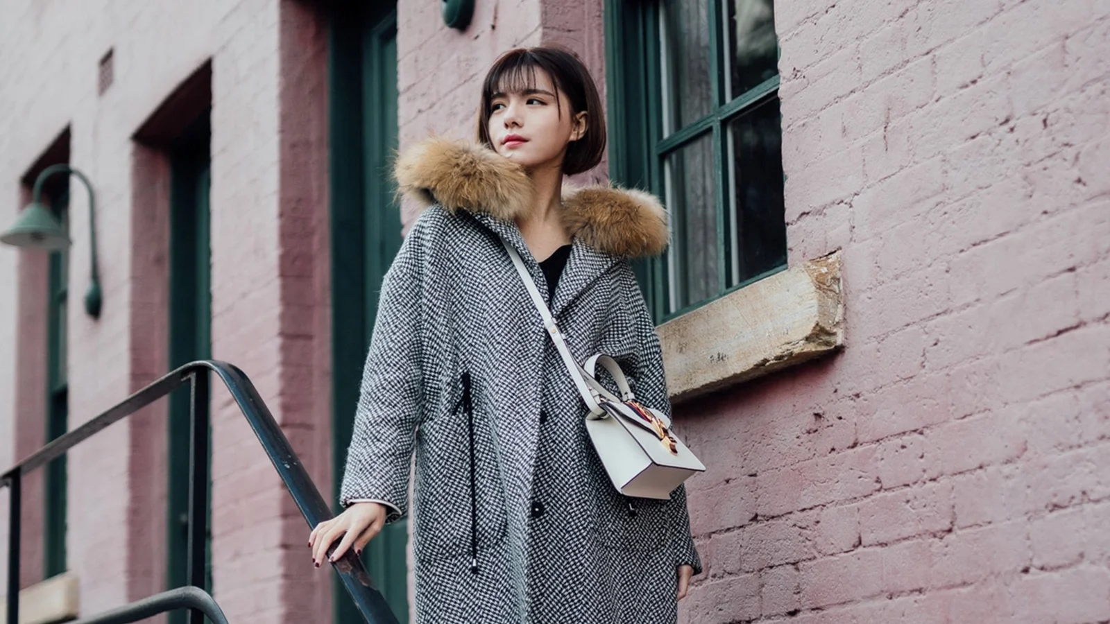 퍼 칼라가 달린 패턴 코트를 입은 성인 여성이 분홍색 벽돌 건물 옆에 서 있습니다.