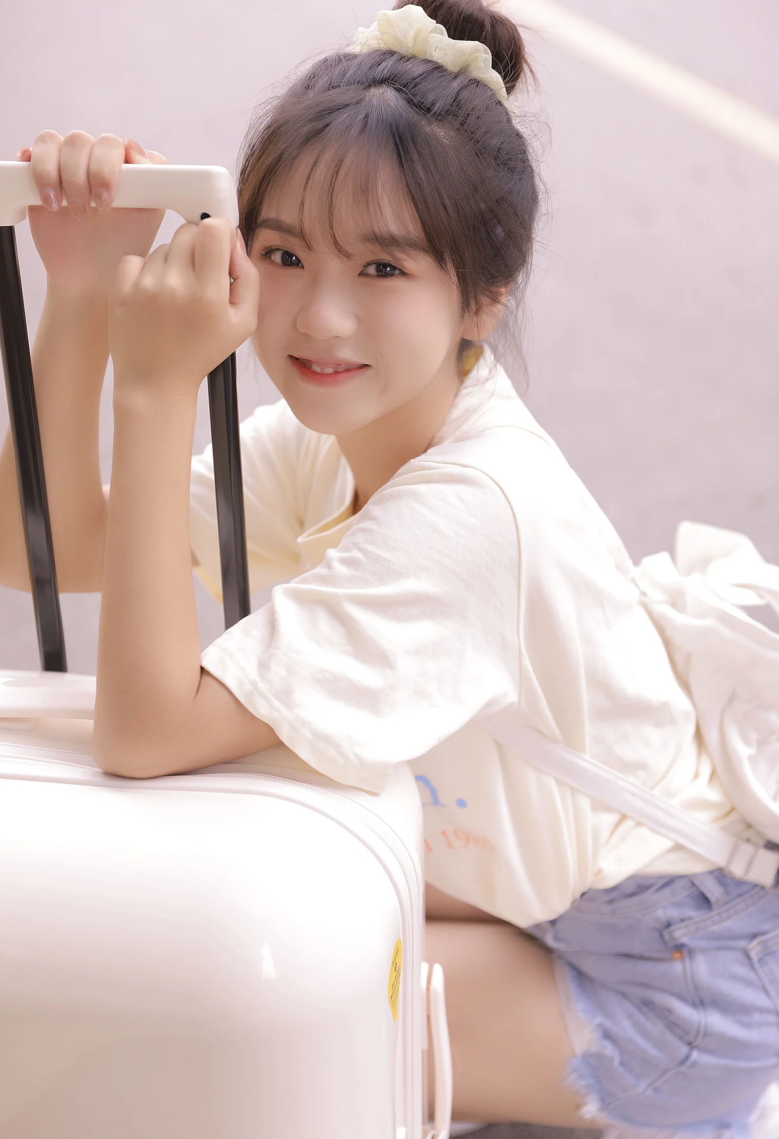 一名盤髮、身穿白色上衣的年輕女子靠在白色行李箱旁看向鏡頭。