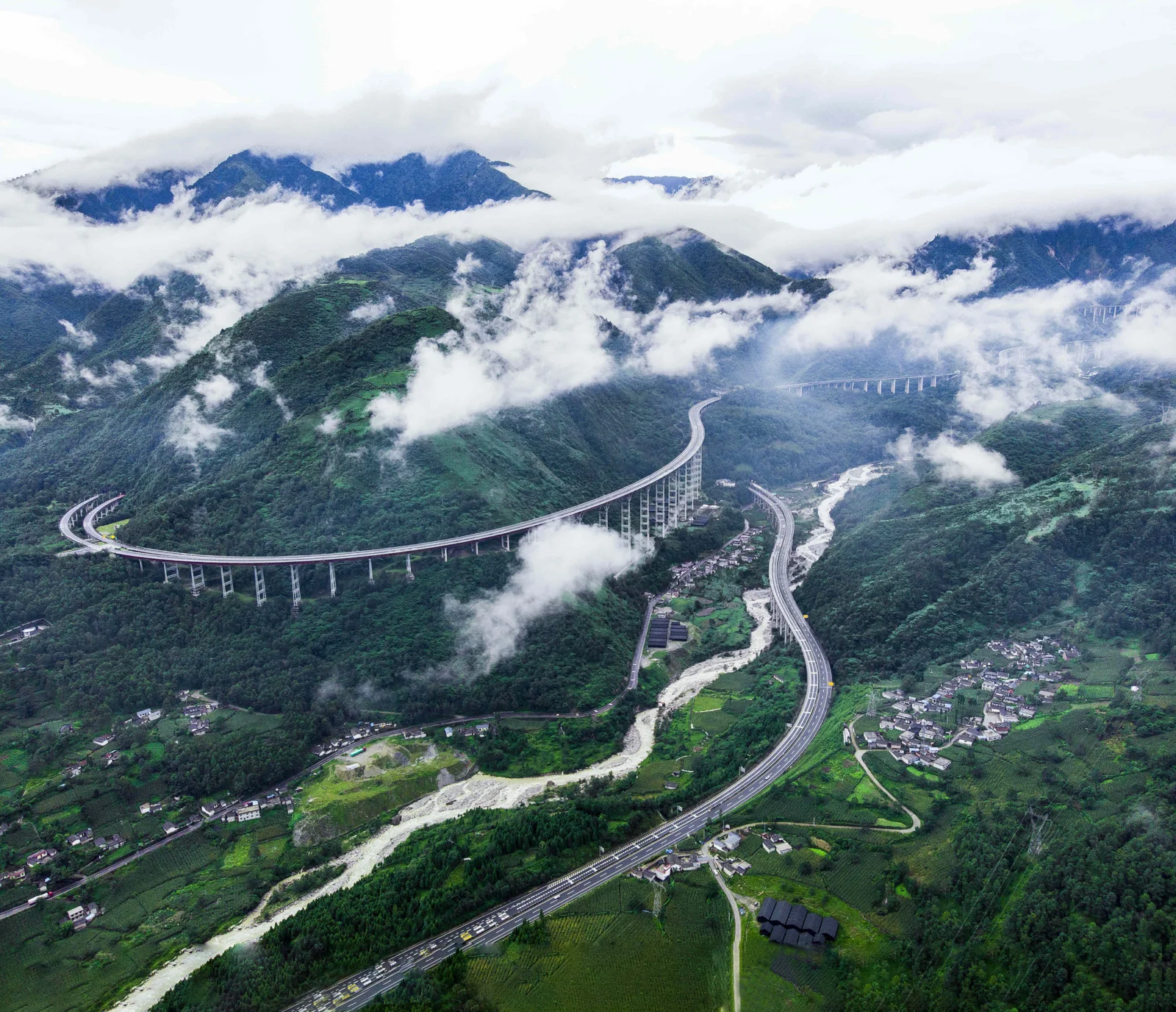 云雾笼罩的绿色山谷中，蜿蜒的高架公路、河流和村落清晰可见。