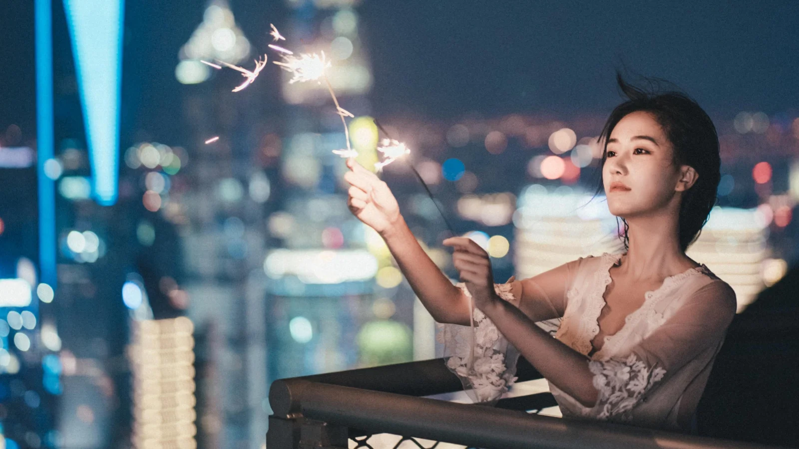 一名成年女性站在城市夜景前的陽台上手持燃燒中的仙女棒。
