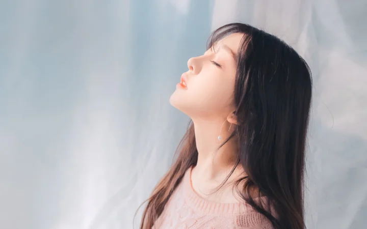 一名长发女性闭眼仰头侧望，身穿浅粉色针织衫，背景为柔和的浅色布幕。