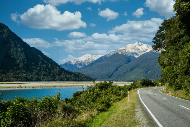 蜿蜒公路沿着河流穿过森林山谷，远处是积雪覆盖的群山和蓝天白云。