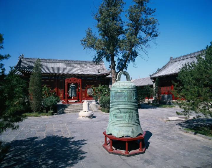 蓝天下的古寺庭院中央摆放着一口大型青铜钟，周围环绕着传统中式建筑和树木。