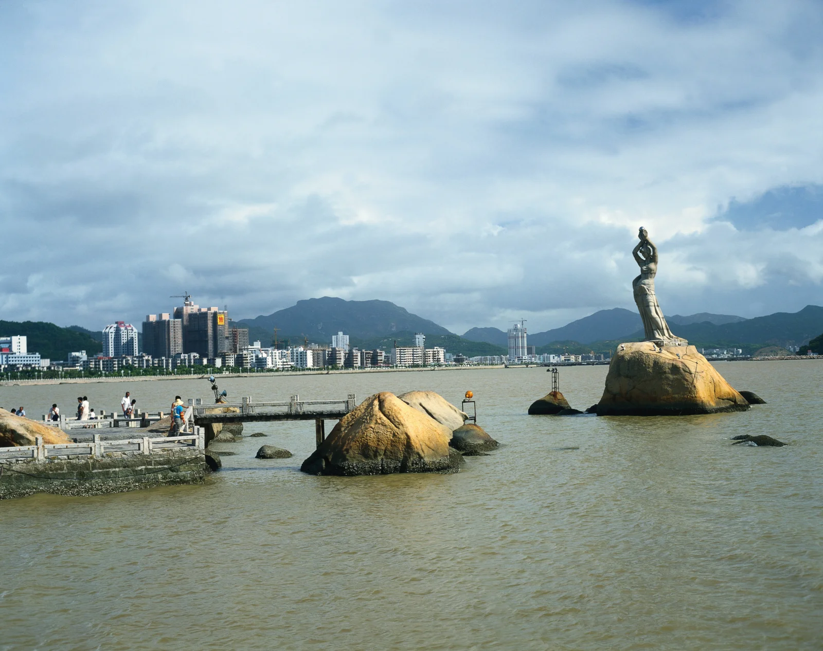 宽阔水面前是一座海滨城市，远处有群山和矗立在巨石上的雕像。
