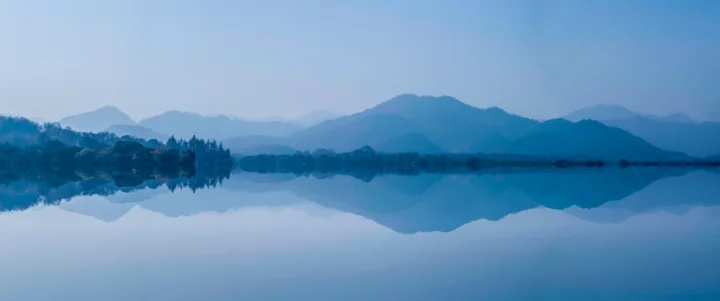 远山、湖水与岸边树木在蓝色雾霭中相映成景，湖面倒映着山峦轮廓。