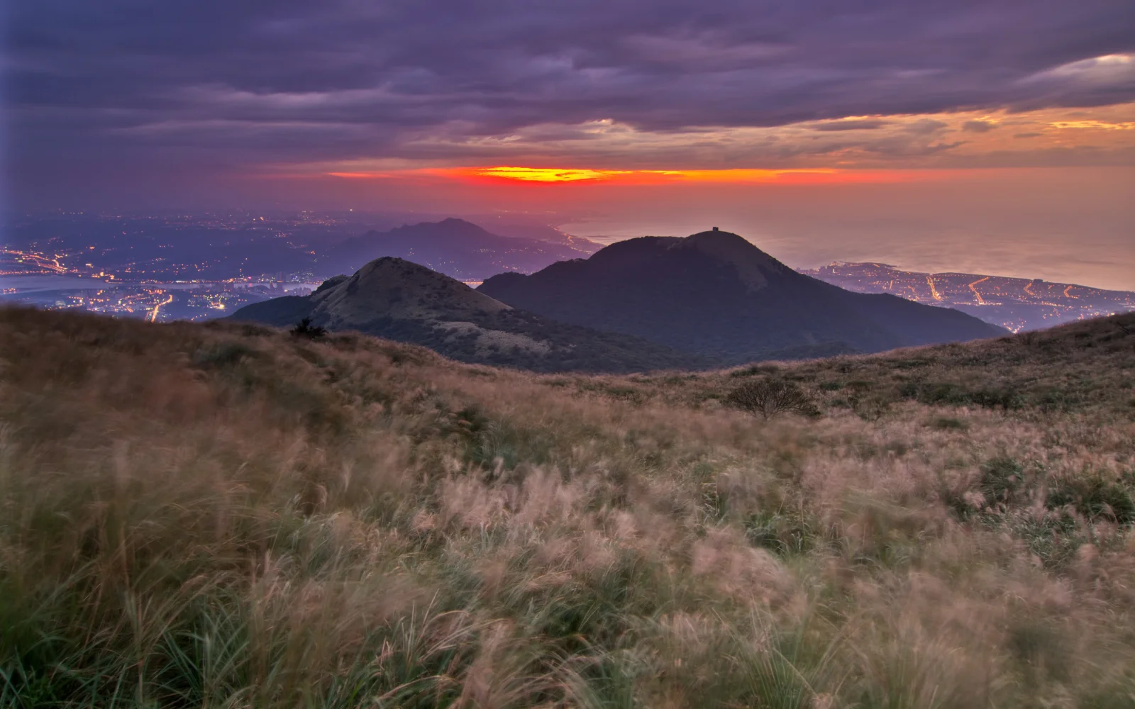 暮色中的山脉、草坡与绚丽日落构成宁静的自然风景，远处可见点点灯光。