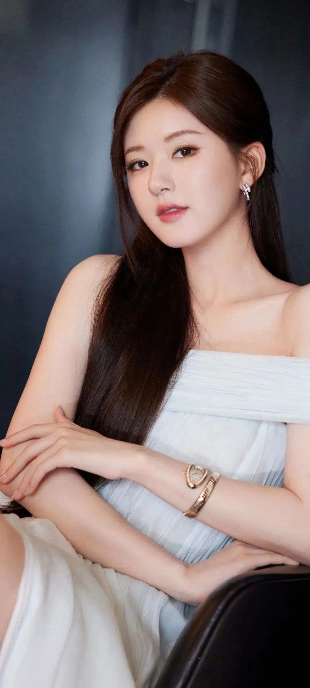 一名长发女性身穿白色露肩连衣裙，坐在深色背景前面对镜头。