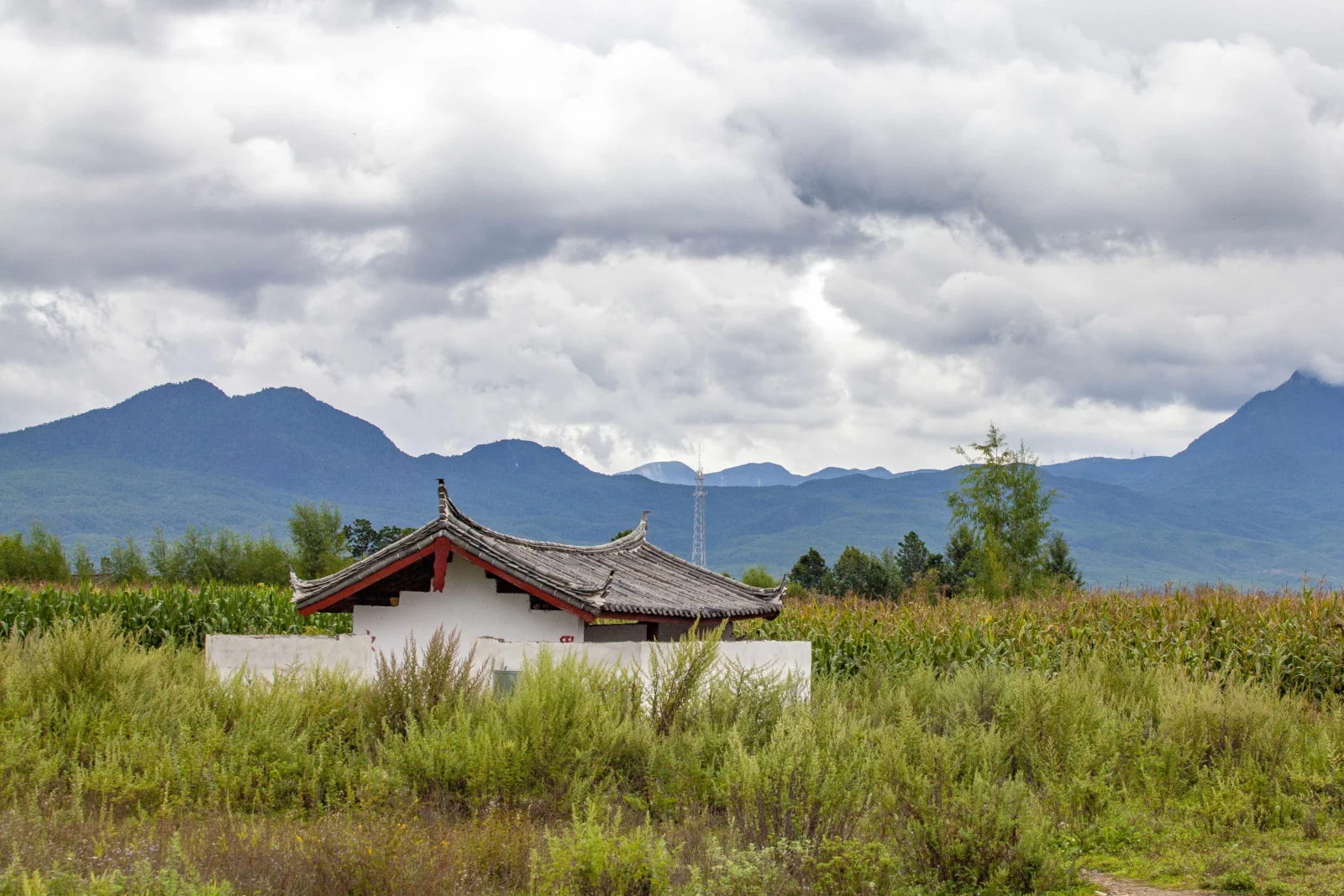 云层低垂的群山、田野与一座传统屋顶建筑构成宁静的自然风景。
