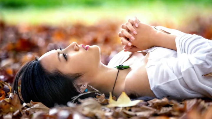 一名女子躺在秋叶上，双手交叠，沐浴在柔和的户外光线中。
