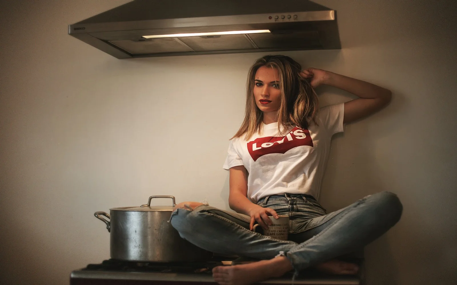 白いTシャツとジーンズを着た成人女性が、金属製の鍋の前で室内に座っています。