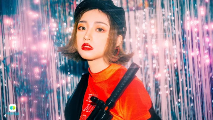 검은색 헤드웨어와 빨간 상의를 착용한 성인 여성이 분홍색과 파란색으로 반짝이는 커튼 앞에 서 있습니다.