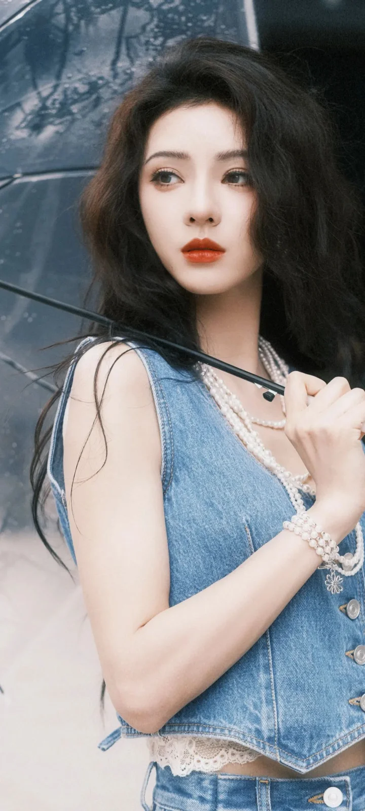 一位长发女性身穿蓝色牛仔服饰，在透明雨伞下拍摄竖幅时尚人像。