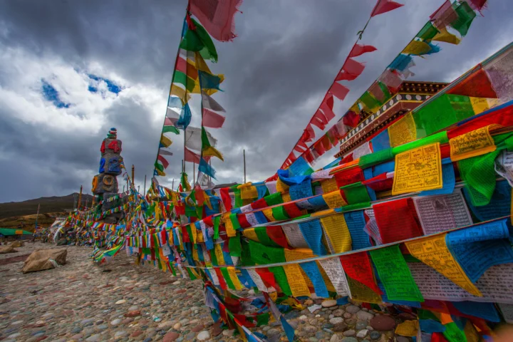 五彩经幡在乌云密布的高原天空下飘扬，远处可见山地与石地。