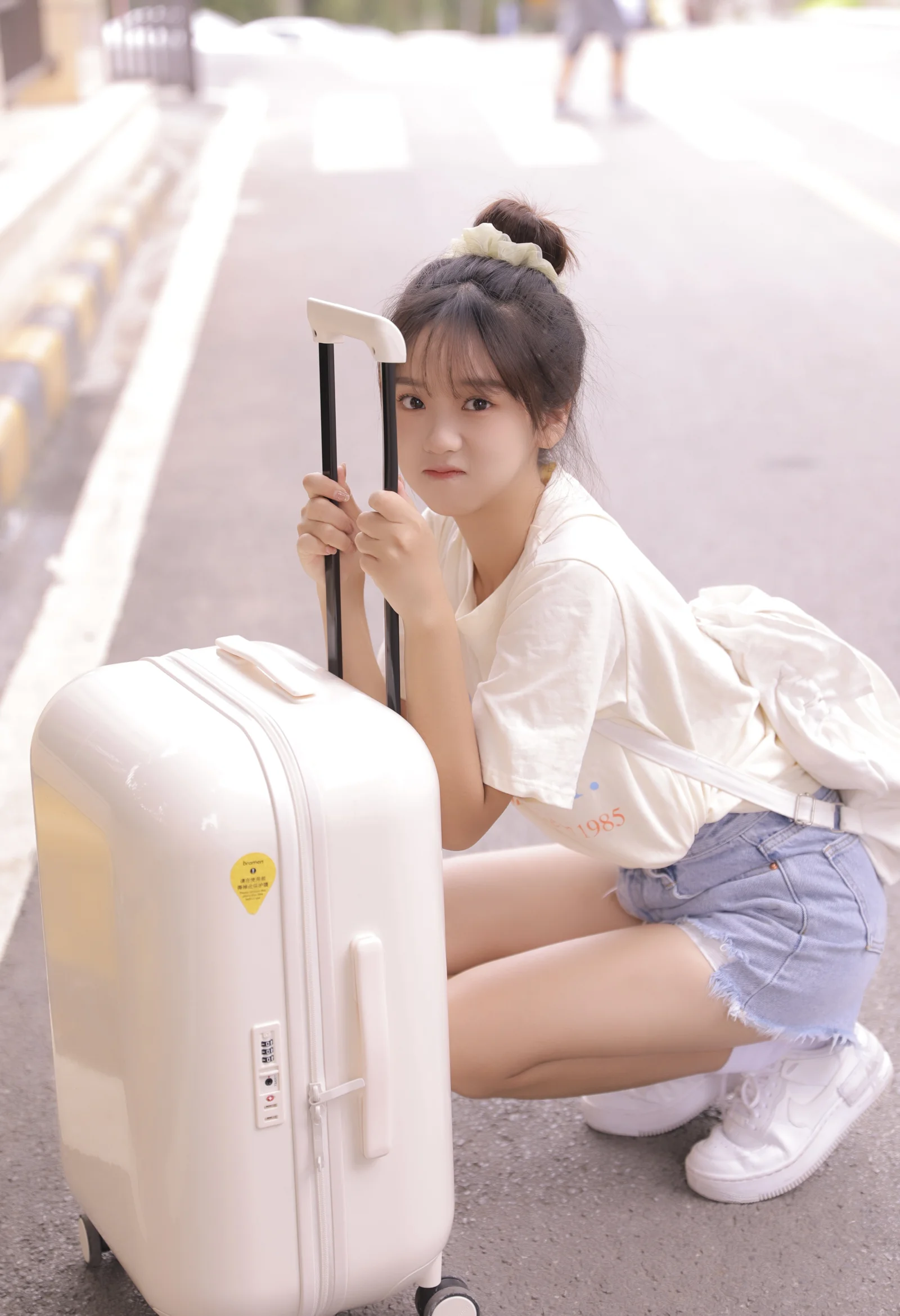 一名年轻女子蹲在街边，手扶着白色行李箱。
