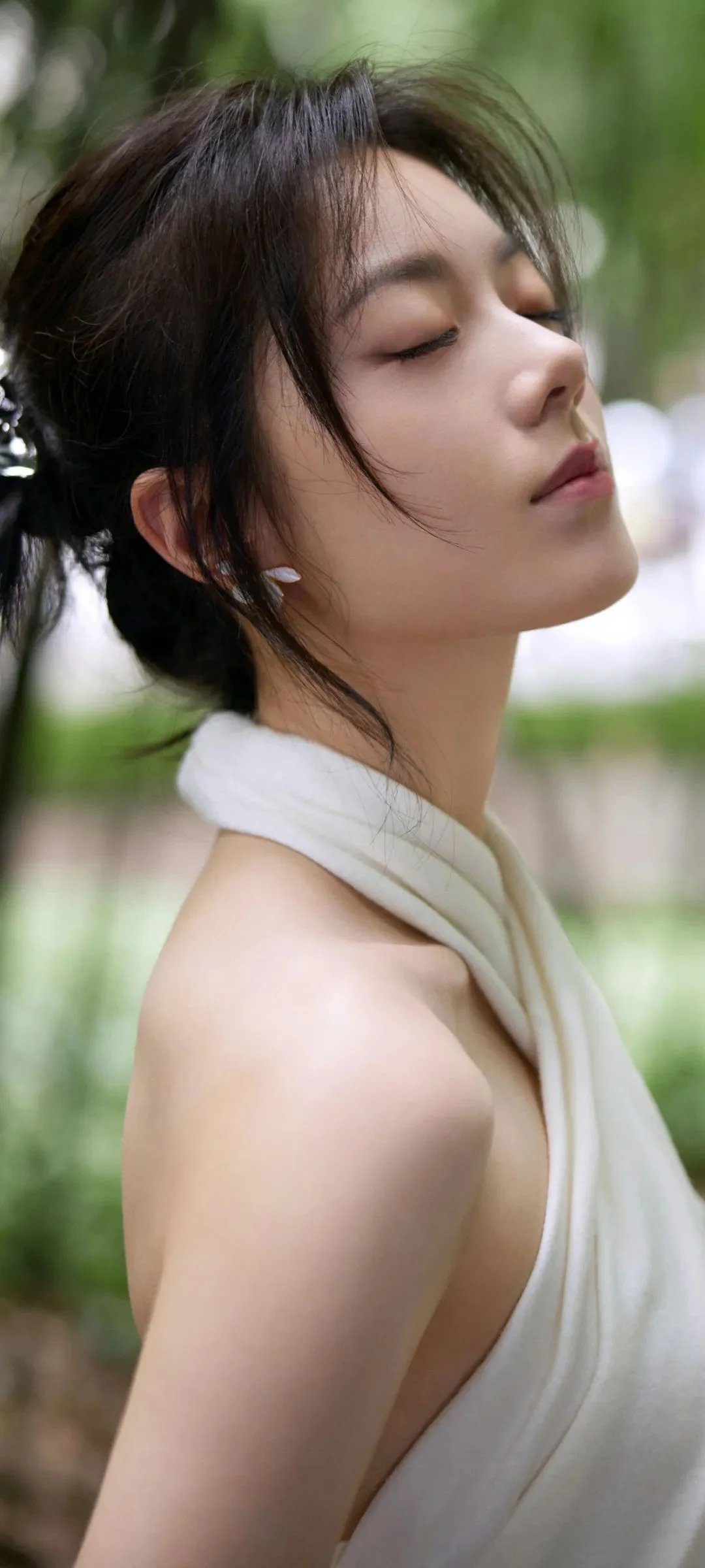 一位女性在户外绿意背景前闭眼侧身，颈间围着浅色围巾。