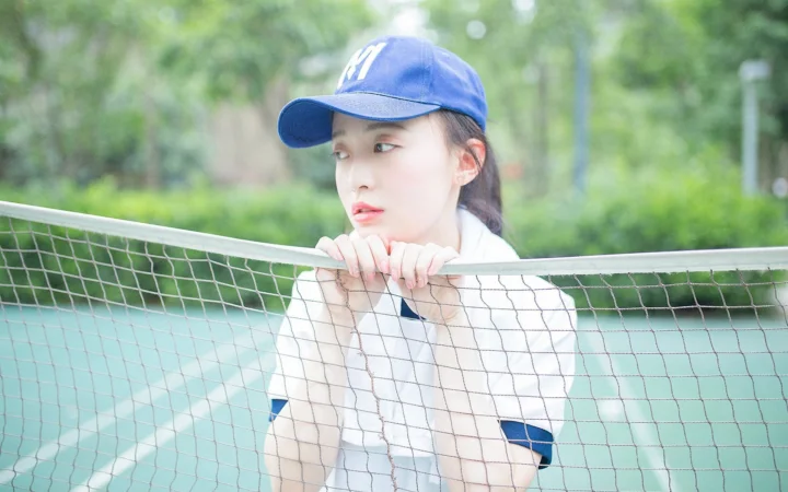 一名戴着蓝色棒球帽的年轻女性站在户外网球网旁。