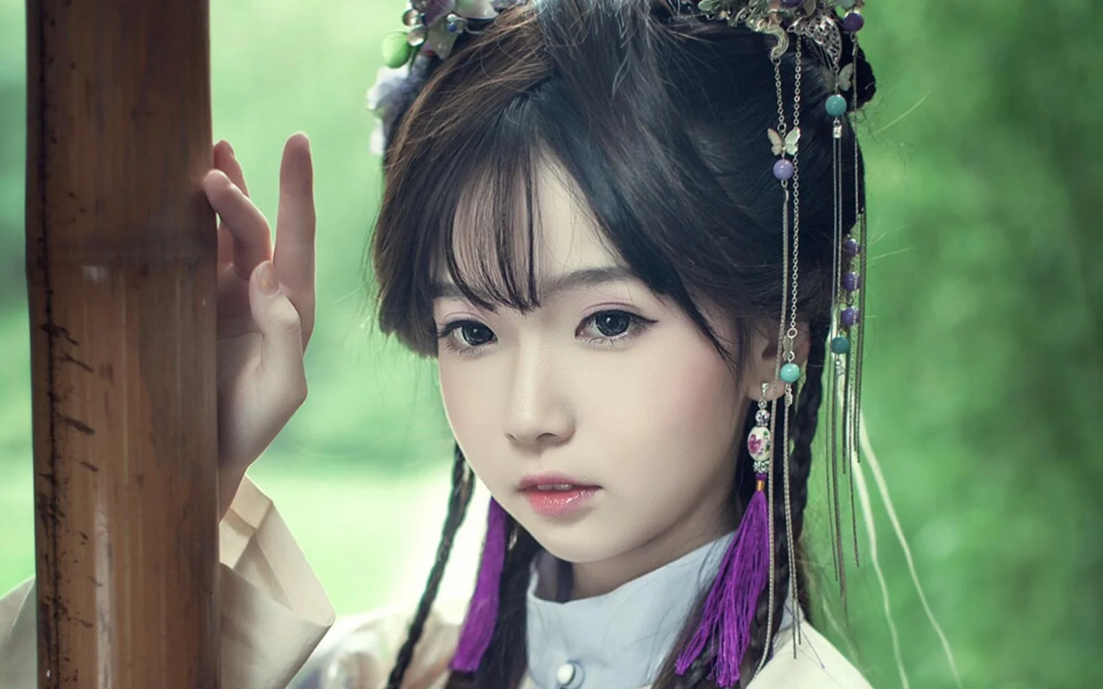 화려한 머리 장식을 한 여성이 부드러운 녹색 배경 앞에서 나무 기둥에 기대고 있습니다.